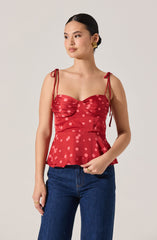 A woman is wearing a sleeveless red top with thin, tied shoulder straps and a sweetheart neckline. The top has a ruched bust area and features a pattern of large pink polka dots. It has a ruffled peplum hem that flares out at the waist. She is also wearing dark blue jeans.