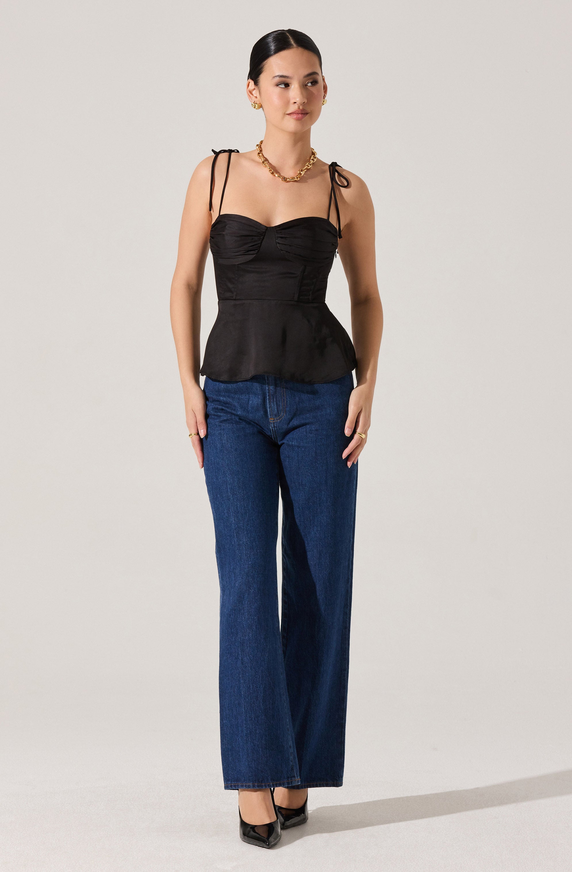 A woman is wearing a black, fitted, sleeveless top with ruched detailing on the bust and thin, tied shoulder straps. She pairs it with high-waisted, wide-leg blue jeans and black pointed-toe high heels. She accessorizes with gold jewelry, including earrings, a necklace, and rings.