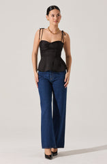 A woman is wearing a black, fitted, sleeveless top with ruched detailing on the bust and thin, tied shoulder straps. She pairs it with high-waisted, wide-leg blue jeans and black pointed-toe high heels. She accessorizes with gold jewelry, including earrings, a necklace, and rings.