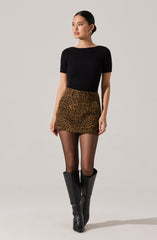 A woman is wearing a fitted black ribbed short-sleeve top, a high-waisted leopard print mini skirt, sheer black tights, and knee-high black leather boots.
