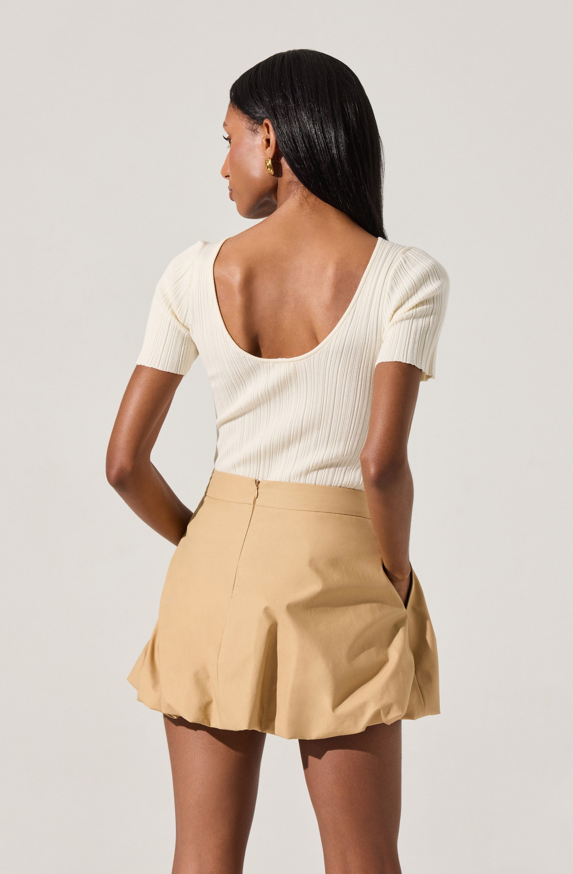 A woman is wearing a cream-colored, ribbed, short-sleeve top with a deep scoop back and puffed sleeves. She pairs it with a high-waisted, beige pleated mini skirt with a slight A-line shape.