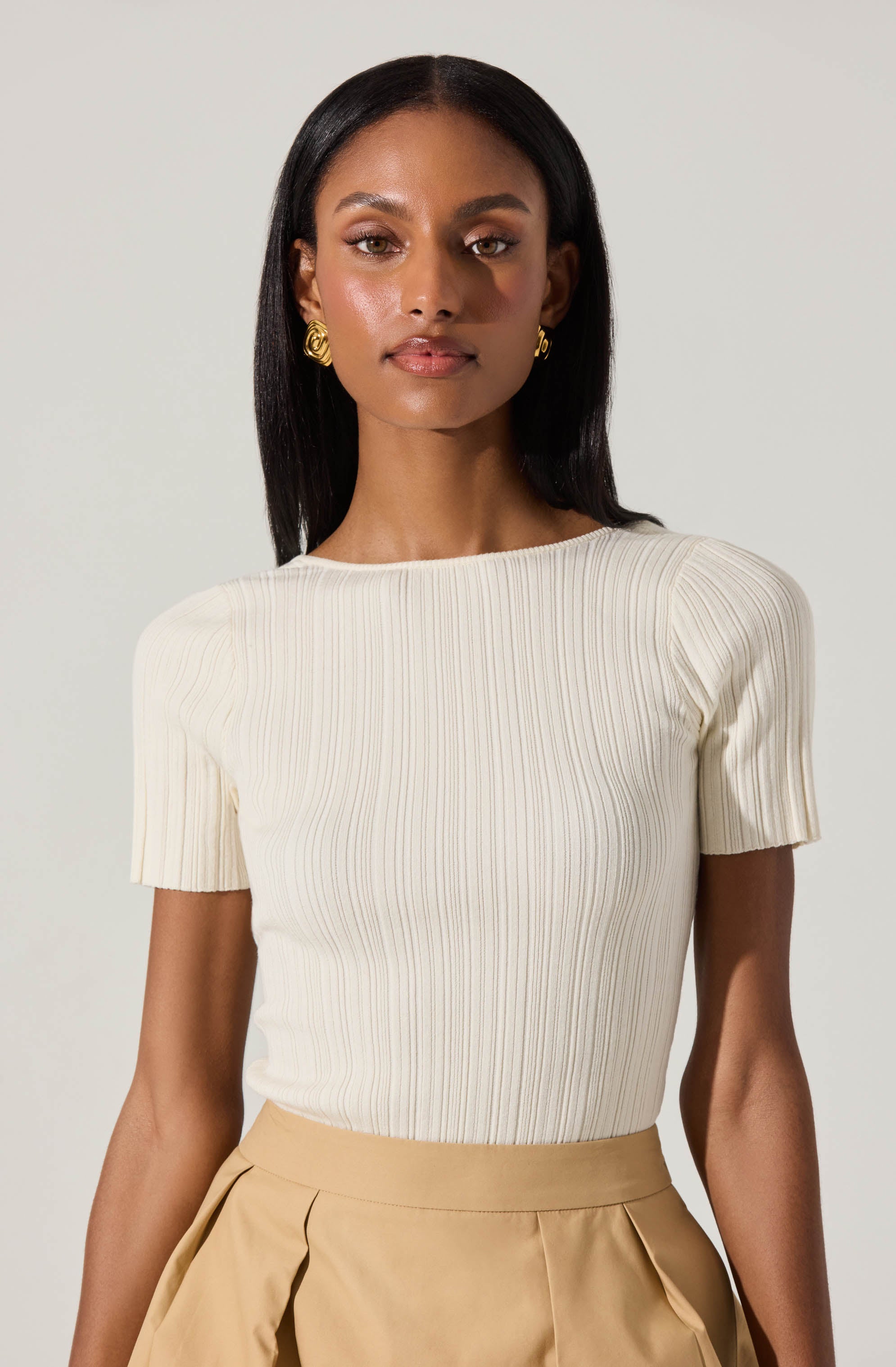 A woman is wearing a short-sleeved, ribbed, cream-colored top with a round neckline. She pairs it with high-waisted, beige pleated trousers.