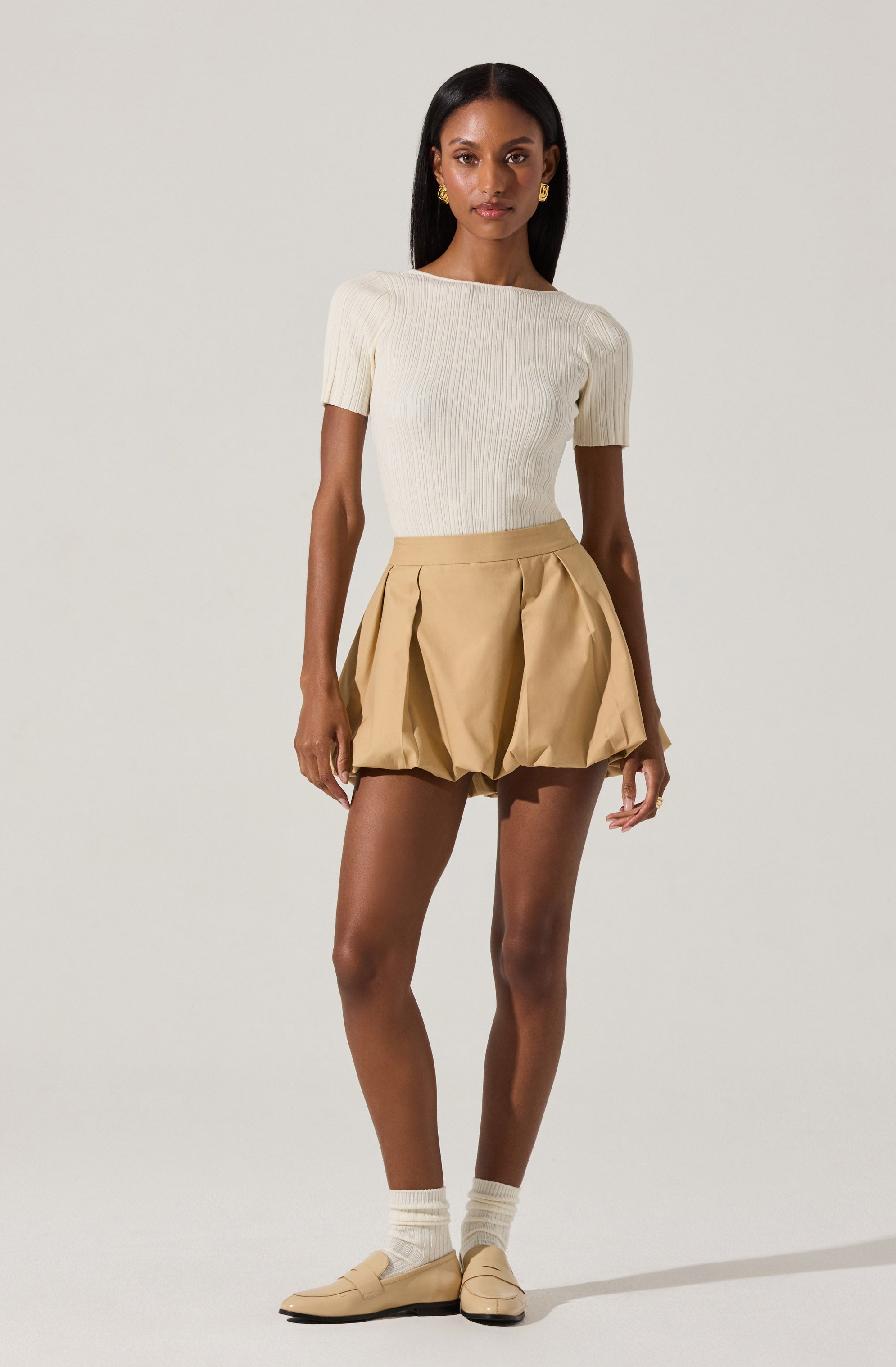 A woman is wearing a fitted, short-sleeved, cream-colored ribbed knit top with a round neckline. She pairs it with high-waisted, tan-colored shorts that have pleats and a puffed, voluminous silhouette. She completes the outfit with beige loafers and cream-colored socks.