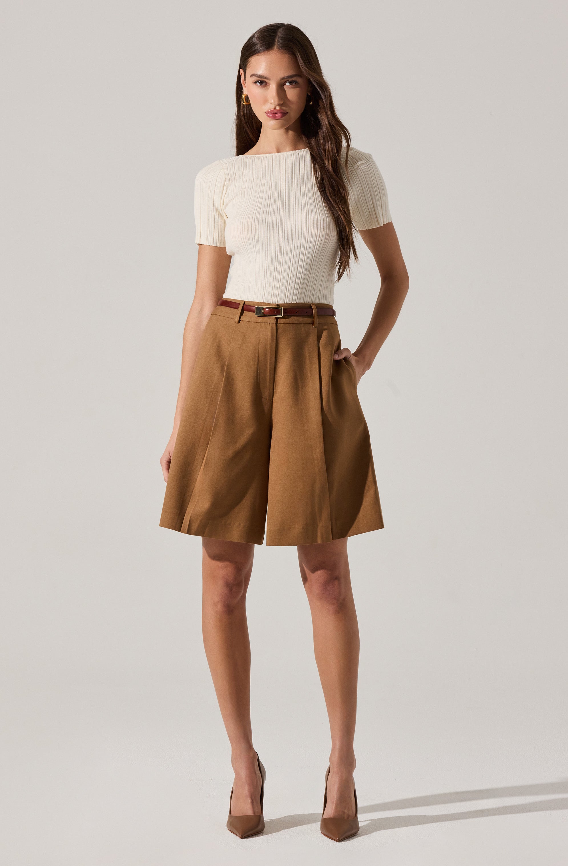 A woman is wearing a short-sleeved, ribbed white knit top paired with wide-leg, high-waisted brown shorts. She accessorizes with a brown belt and brown pointed-toe high heels.