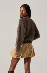 A woman is wearing a loose, long-sleeved, fuzzy, dark brown sweater and a high-waisted, beige, pleated mini skirt.