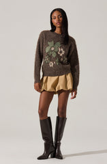A woman is wearing a dark brown fuzzy sweater with a large floral pattern in light green, white, and beige on the front. She has on a beige, puffy, knee-length skirt with gathered fabric, and black knee-high boots with a pointed toe and a block heel.