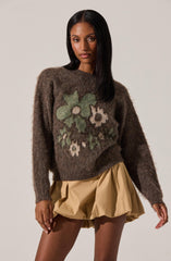 A woman is wearing a cozy, oversized brown sweater with large, abstract floral patterns in shades of green, white, and beige on the front. She pairs it with beige, voluminous, bubble-style shorts.