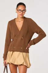 A woman is wearing a brown, double-breasted knit cardigan with a V-neckline, featuring decorative gold buttons and textured vertical stitching on the front. She pairs it with a beige, bubble-hem skirt.