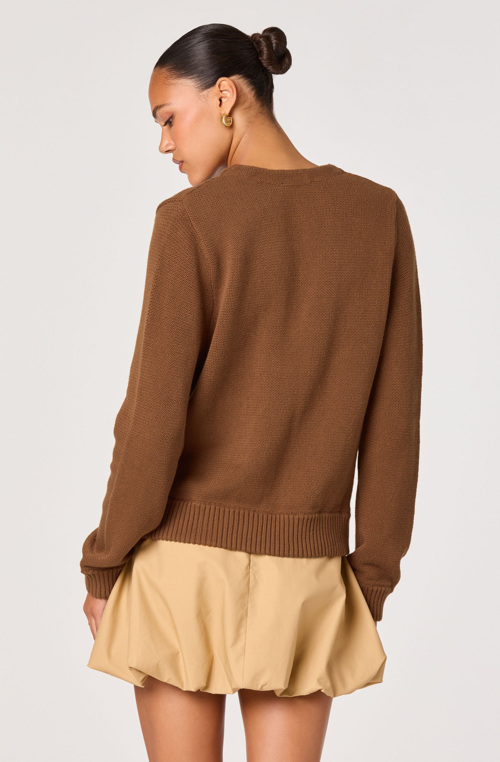 A woman is wearing a brown, long-sleeved knitted sweater with ribbed cuffs and hem. The sweater has a simple, round neckline and a textured pattern. She pairs it with a beige, puffed skirt that has a gathered, voluminous design. The woman has dark hair styled in a sleek bun and is accessorized with gold hoop earrings.