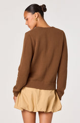 A woman is wearing a brown, long-sleeved knitted sweater with ribbed cuffs and hem. The sweater has a simple, round neckline and a textured pattern. She pairs it with a beige, puffed skirt that has a gathered, voluminous design. The woman has dark hair styled in a sleek bun and is accessorized with gold hoop earrings.