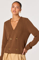 A woman is wearing a brown knitted cardigan with a deep V-neckline, featuring decorative gold buttons arranged in two vertical rows along the front. The cardigan has textured vertical and horizontal patterns around the shoulders and down the front. The sleeves are long with ribbed cuffs.
