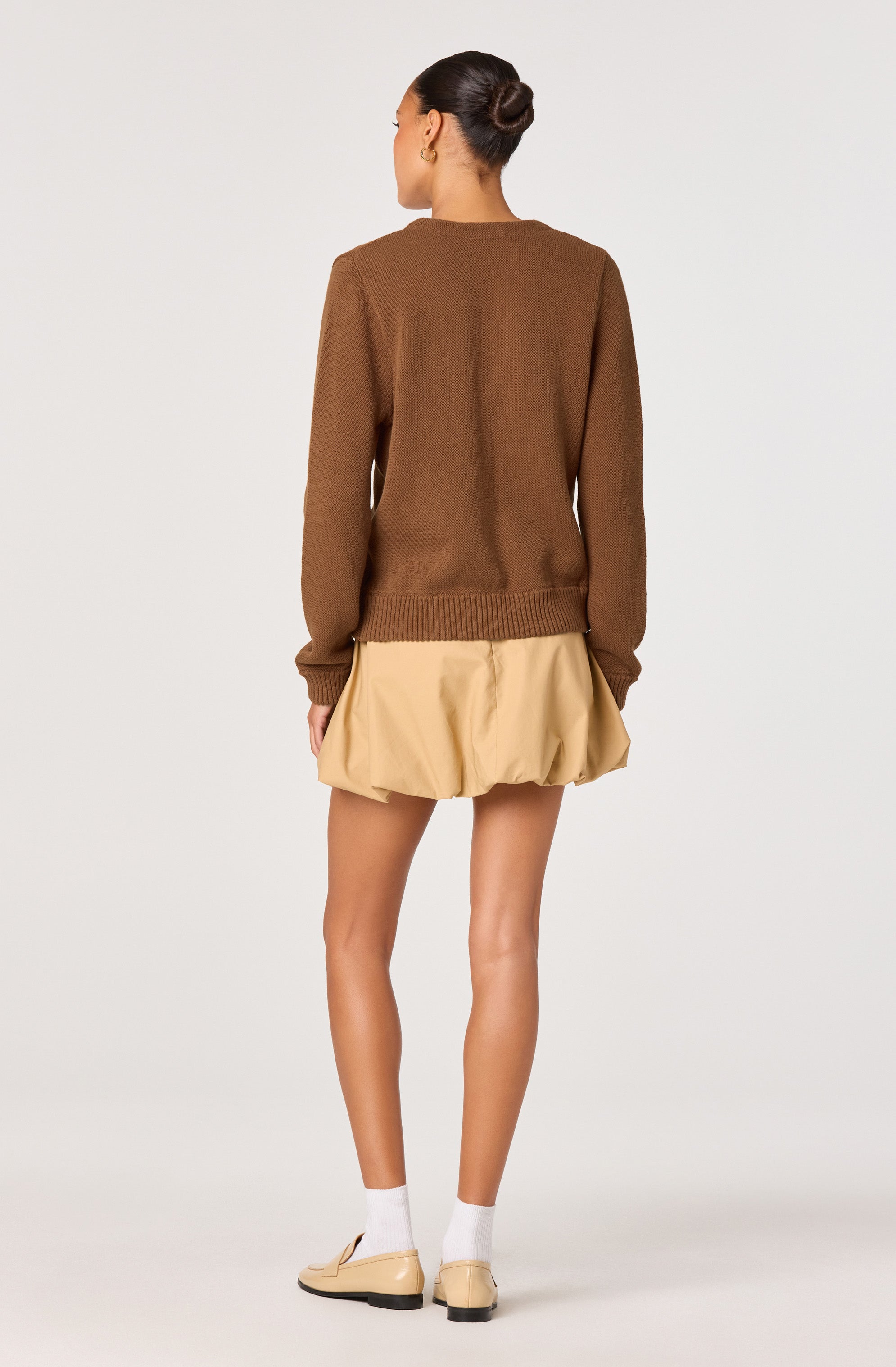 A woman is wearing a brown long-sleeve sweater with ribbed cuffs and hem, paired with a beige, gathered, bubble-style skirt that has a loose, puffy appearance. She has her hair styled in a neat bun and is wearing white socks and beige flat shoes.