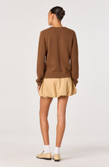 A woman is wearing a brown long-sleeve sweater with ribbed cuffs and hem, paired with a beige, gathered, bubble-style skirt that has a loose, puffy appearance. She has her hair styled in a neat bun and is wearing white socks and beige flat shoes.