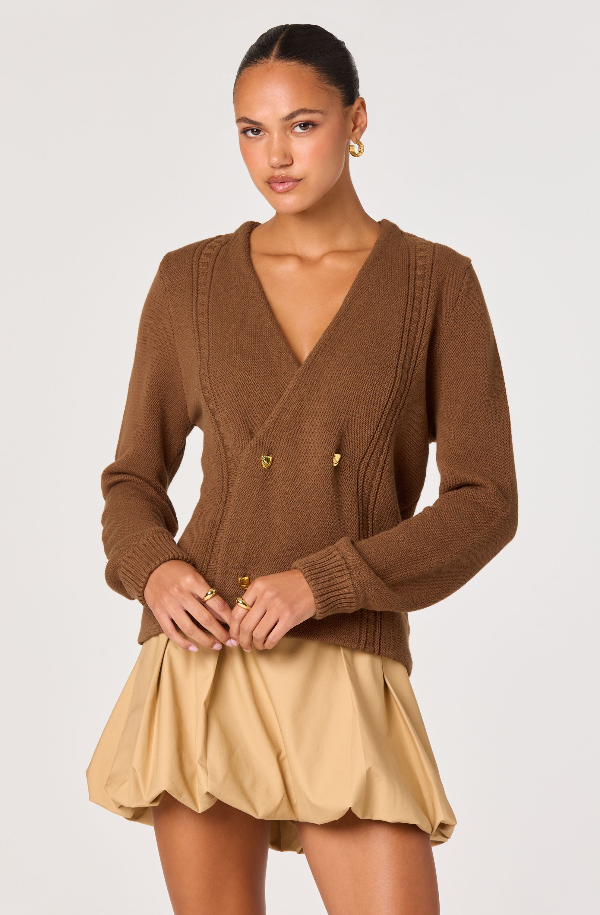 A woman is wearing a brown knit sweater with a V-neckline and decorative stitching along the shoulders and front. The sweater has long sleeves with ribbed cuffs and gold button accents at the front. She pairs it with beige, voluminous, ruched shorts that have a gathered, bubble-like appearance.