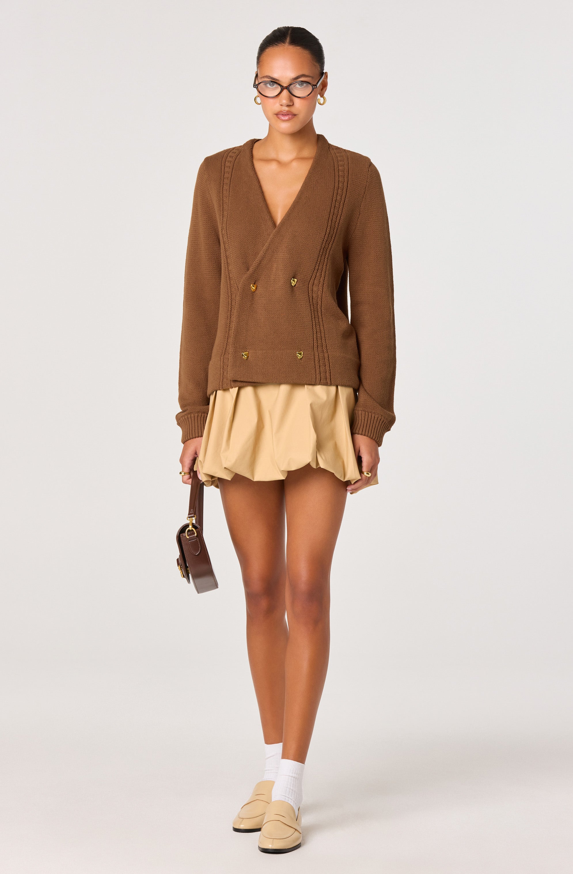 A woman is wearing a brown, double-breasted knit cardigan with gold buttons, featuring a V-neckline and long sleeves. She pairs it with a beige, ruffled, above-the-knee skirt. She accessorizes with glasses, gold hoop earrings, a small brown handbag, white socks, and beige shoes.