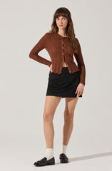 A woman is wearing a brown, long-sleeved, knit cardigan with a textured pattern, including a lace detail on the collar. She has paired it with a black mini skirt, cream-colored socks, and black loafers with buckle accents.