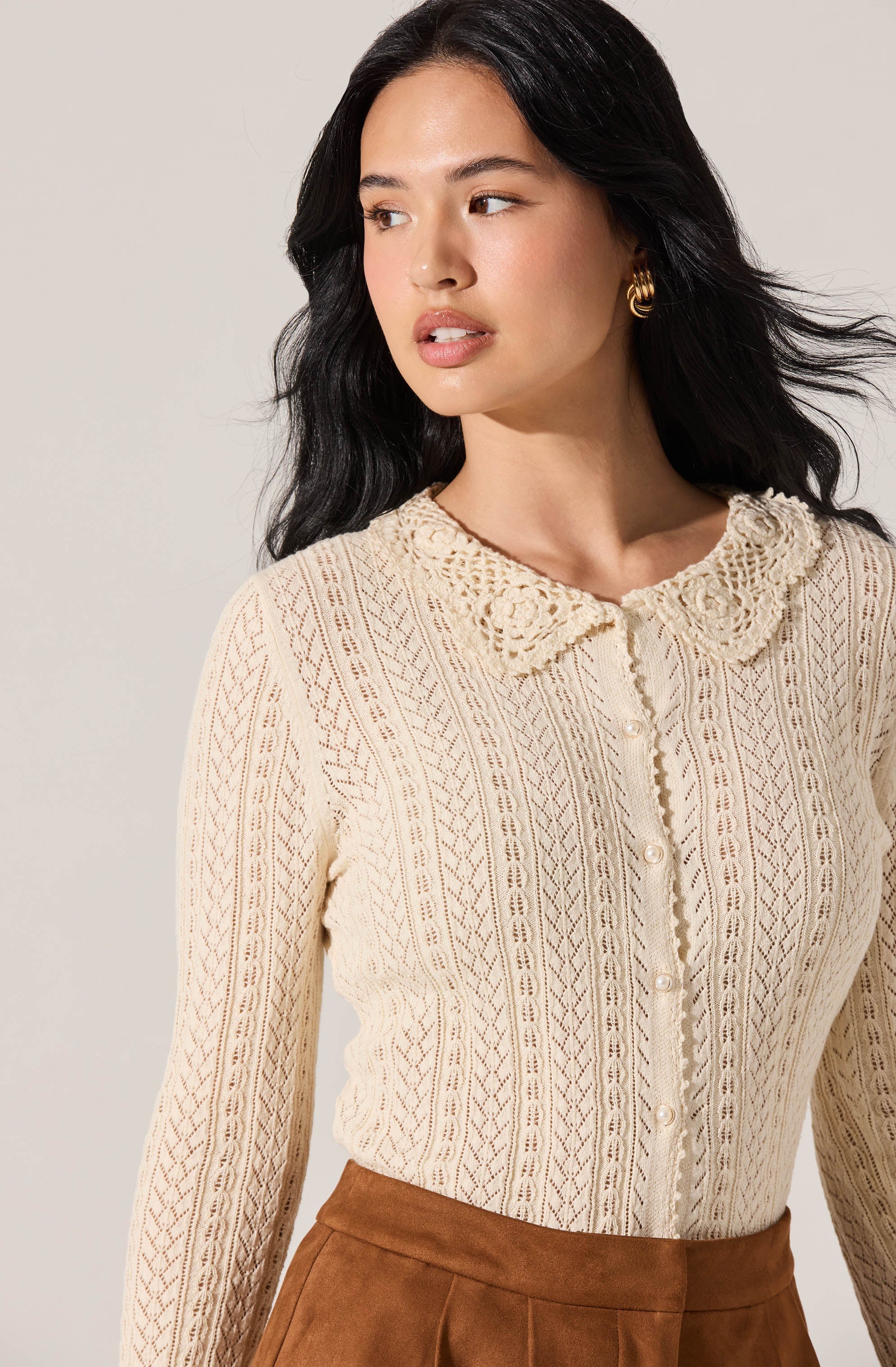 A woman wearing a long-sleeved, cream-colored knit sweater with an intricate openwork pattern. The sweater has a rounded, lace-trimmed collar and button-down front. She pairs it with high-waisted brown pants.
