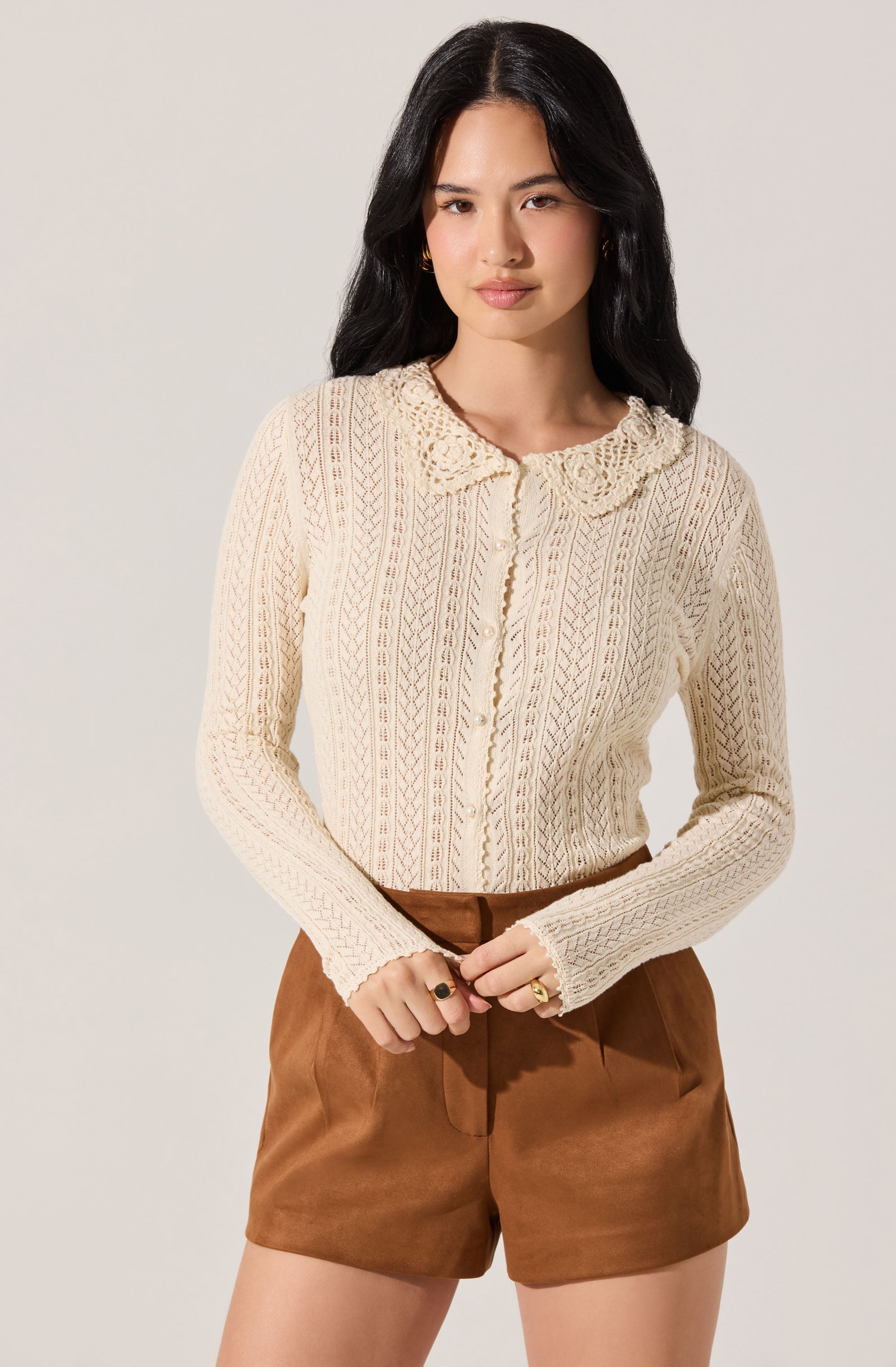 A woman is wearing a cream-colored, long-sleeved, knitted sweater with a textured pattern and a scalloped lace collar. She pairs it with high-waisted brown shorts made of a soft, suede-like material.