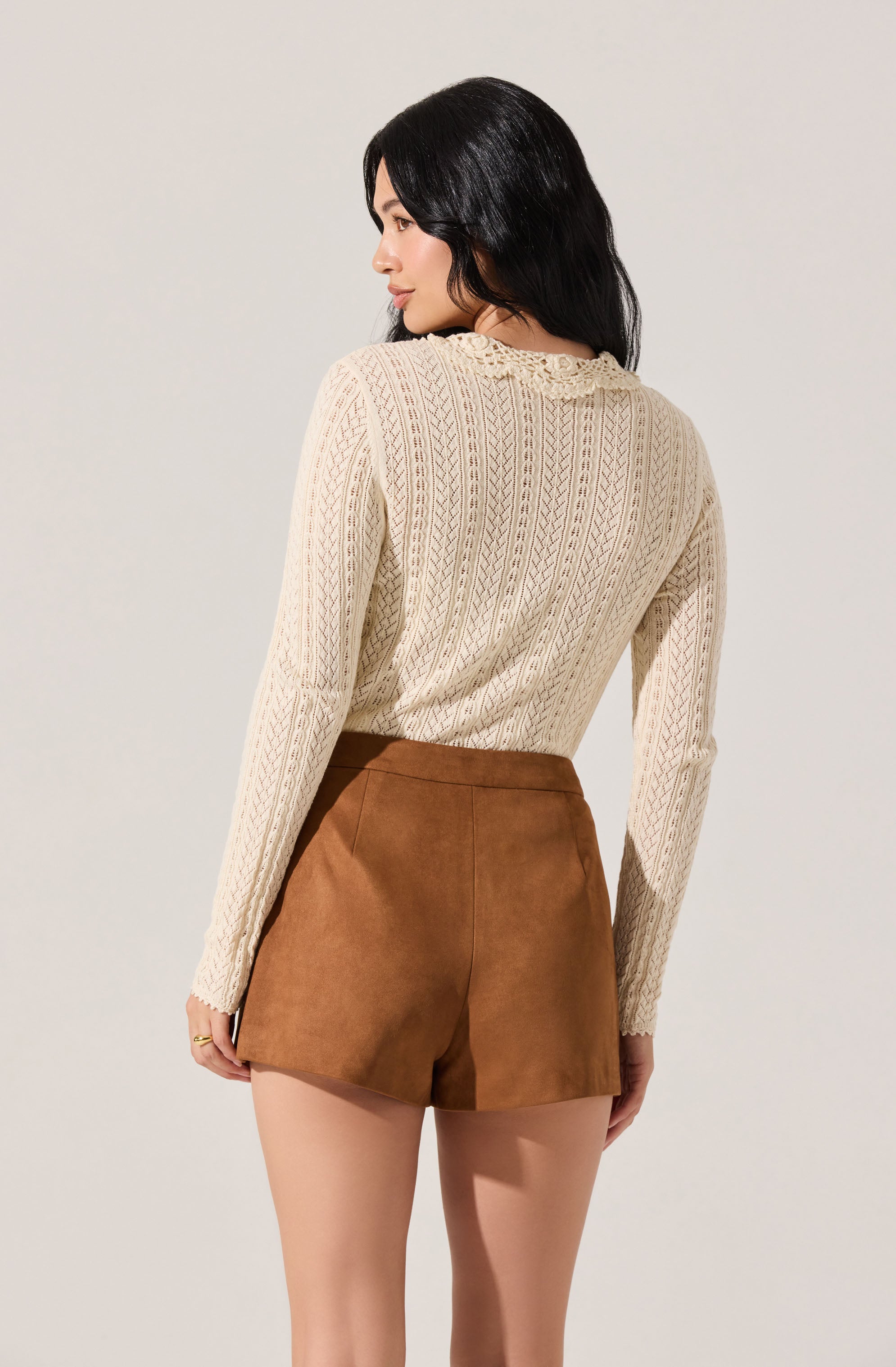A woman is wearing a cream-colored, long-sleeve sweater with a textured, cable-knit pattern. She pairs it with high-waisted, brown faux suede shorts.