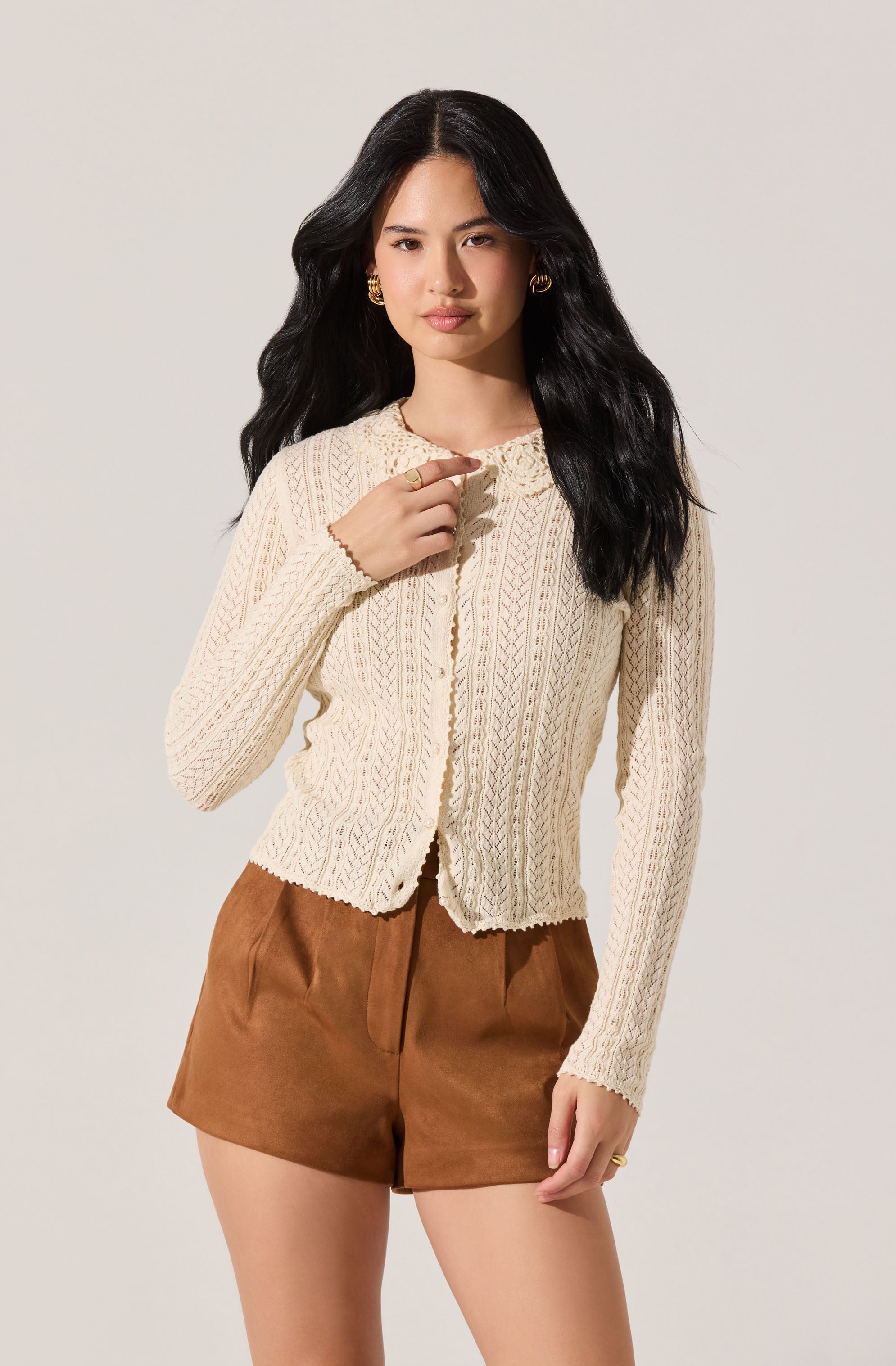 A woman is wearing a cream-colored, long-sleeved, button-up knit cardigan with a textured, cable-knit pattern. She has paired it with high-waisted, brown shorts made of a soft, suede-like material.