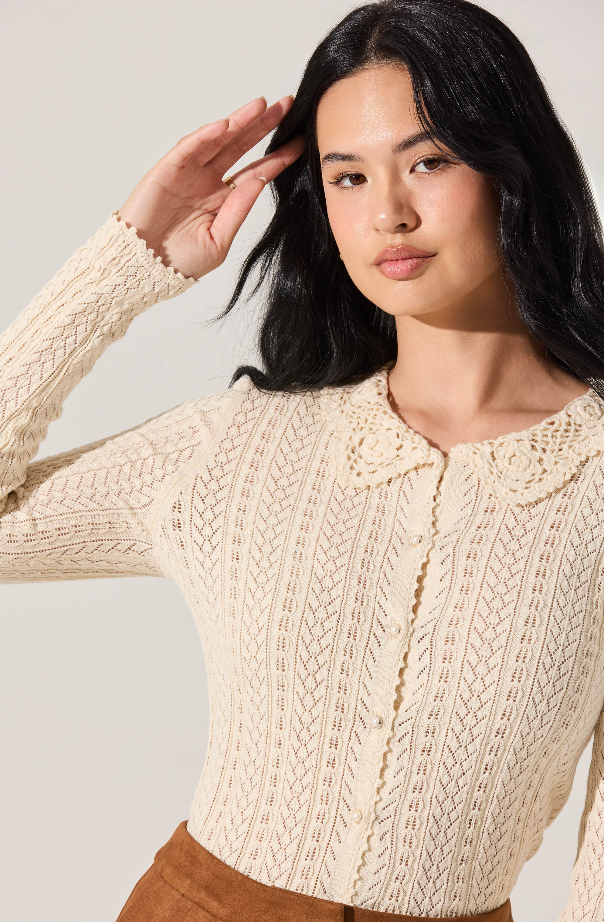 A woman is wearing a cream-colored, long-sleeve, knit cardigan with intricate lace and crochet patterns. The cardigan features a scalloped lace collar and pearl-like buttons down the front.
