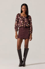 A woman is wearing a long-sleeved, loose-fitting blouse with a floral pattern in shades of pink, cream, and brown on a dark background. The blouse has a square neckline and slightly puffed sleeves. She pairs it with a high-waisted, burgundy mini skirt made of a smooth fabric, featuring a side zipper and a decorative chain detail hanging from the waist. She completes her outfit with tall, black leather knee-high boots.