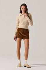 A young woman is wearing a cream-colored, long-sleeve, knit sweater with a textured diamond pattern and a collared neckline. She has paired it with a brown corduroy mini skirt and cream-colored socks, along with beige loafers.