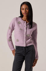 A woman is wearing a light purple, fuzzy cardigan decorated with large, shiny, rhinestone embellishments scattered across the front, sleeves, and shoulders. The cardigan has a round neckline and button closure down the front. She pairs it with high-waisted, dark gray jeans.