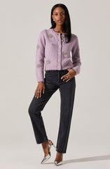 A woman is wearing a light purple, fuzzy cardigan with decorative silver embellishments on the front, sleeves, and shoulders. The cardigan has a round neckline and buttons down the front. She is pairing it with high-waisted black jeans and silver high-heeled shoes.