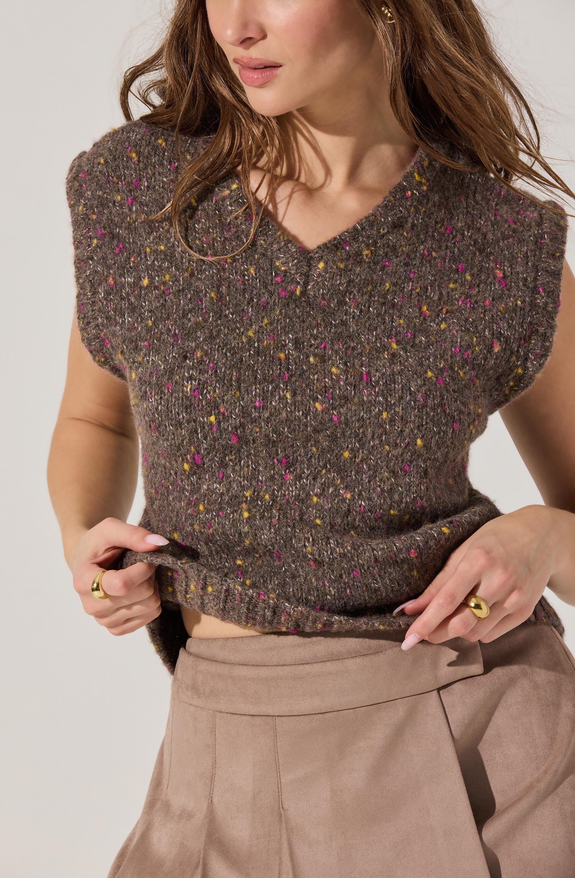 A woman is wearing a sleeveless, V-neck knit top in a brown tweed pattern with multicolored specks of pink, yellow, and purple. The top appears soft and slightly loose-fitting. She is also wearing beige high-waisted pants with a pleated front.