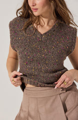 A woman is wearing a sleeveless, V-neck knit top in a brown tweed pattern with multicolored specks of pink, yellow, and purple. The top appears soft and slightly loose-fitting. She is also wearing beige high-waisted pants with a pleated front.