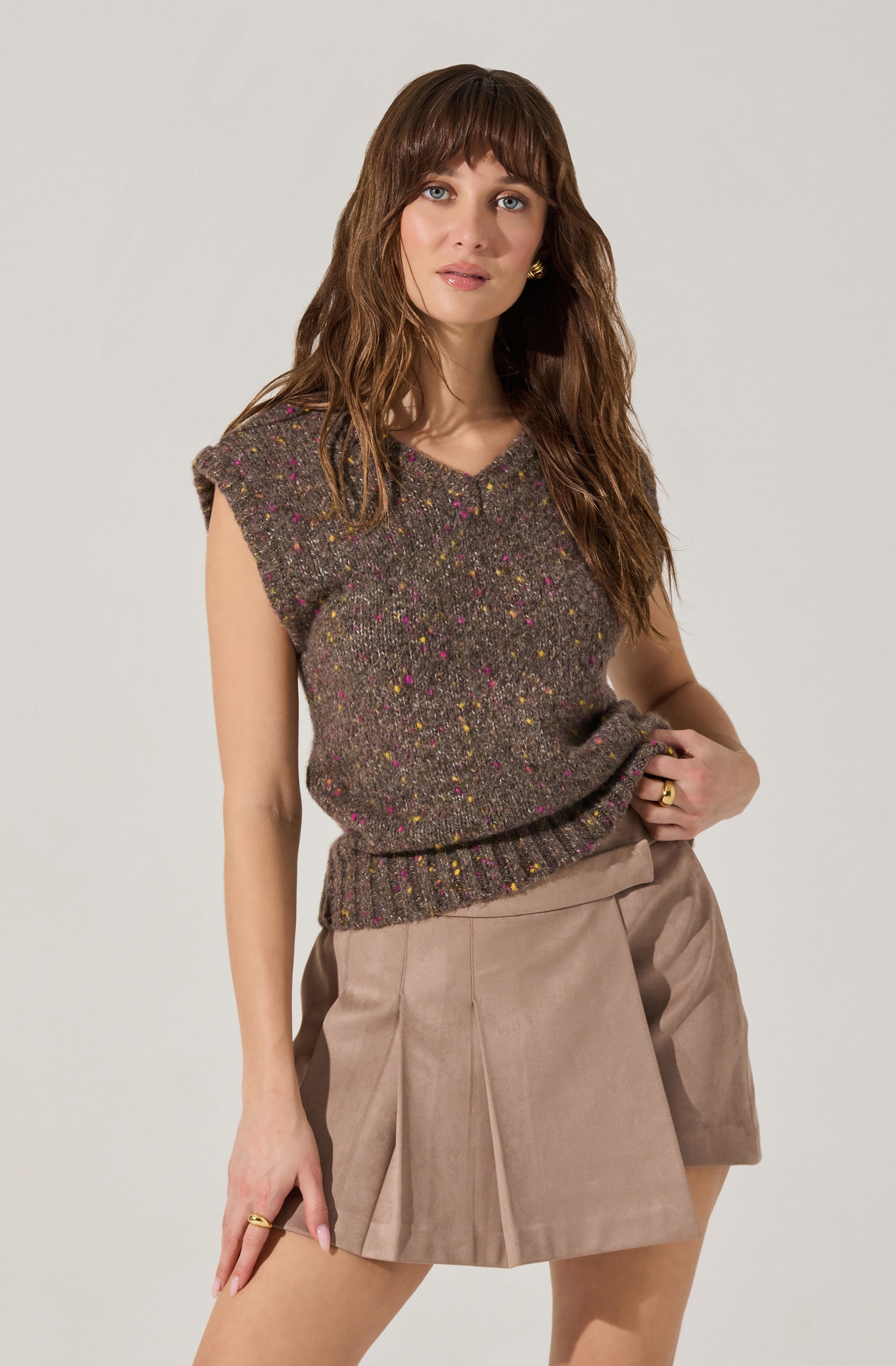 A woman is wearing a sleeveless, knitted sweater in a dark speckled pattern with pink, yellow, and green flecks. She pairs it with beige high-waisted shorts that have pleats in the front. She has wavy brown hair with bangs and is accessorized with gold earrings and rings.
