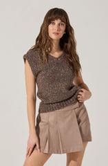 A woman is wearing a sleeveless, knitted sweater in a dark speckled pattern with pink, yellow, and green flecks. She pairs it with beige high-waisted shorts that have pleats in the front. She has wavy brown hair with bangs and is accessorized with gold earrings and rings.