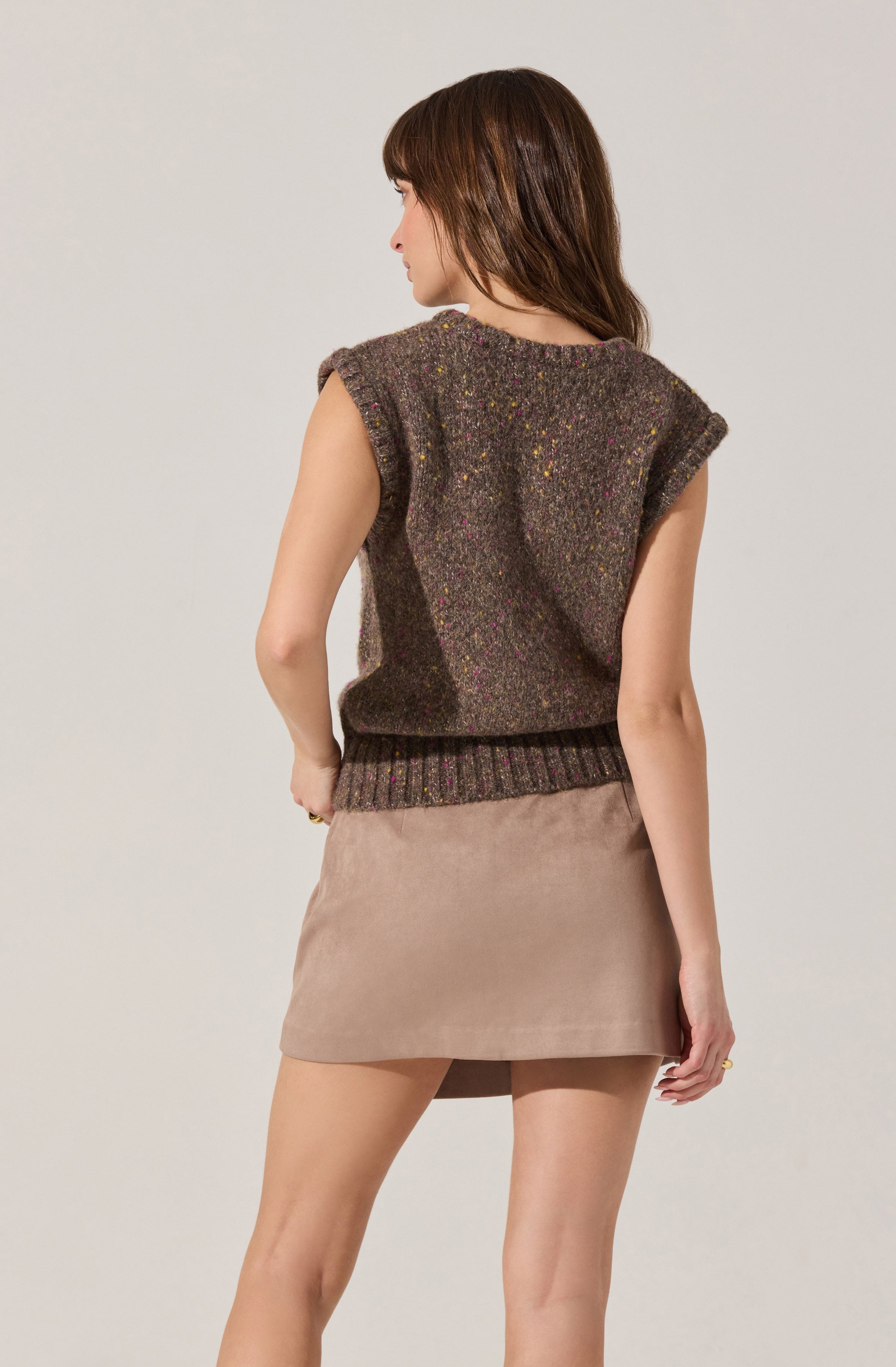 A woman is wearing a sleeveless, oversized, dark brown knit sweater with colorful speckles throughout. The sweater has ribbed detailing at the hem, and the shoulder area is rolled up. She pairs it with a beige suede mini skirt.