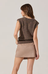 A woman is wearing a sleeveless, oversized, dark brown knit sweater with colorful speckles throughout. The sweater has ribbed detailing at the hem, and the shoulder area is rolled up. She pairs it with a beige suede mini skirt.