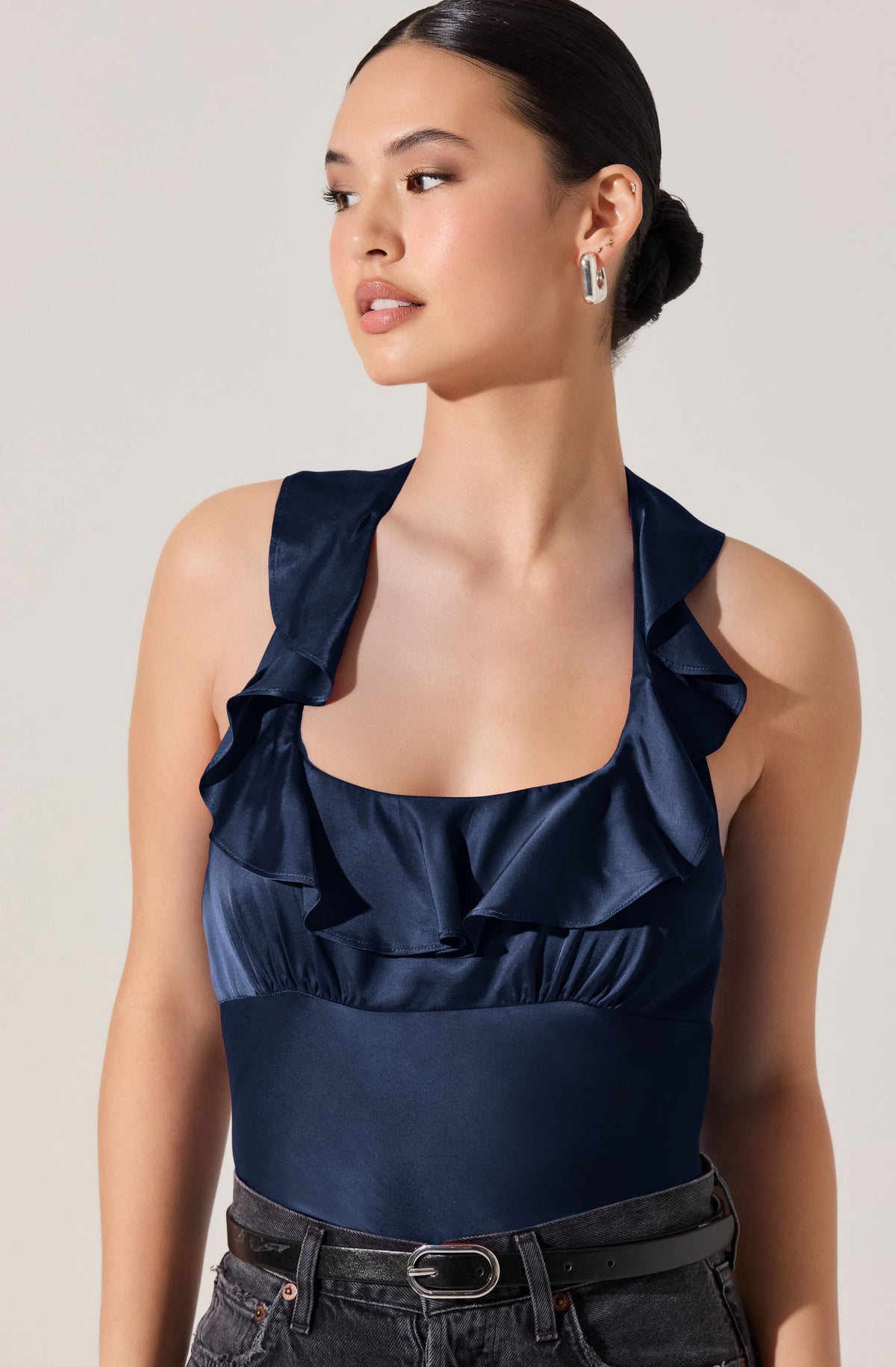 A woman is wearing a navy blue sleeveless top made of shiny satin fabric. The top features ruffled straps and a ruffled neckline. She is also wearing dark jeans with a black belt that has a metallic buckle. The woman has her hair styled in a low bun and is accessorized with large, silver hoop earrings.