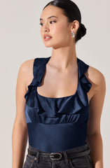 A woman is wearing a navy blue sleeveless top made of shiny satin fabric. The top features ruffled straps and a ruffled neckline. She is also wearing dark jeans with a black belt that has a metallic buckle. The woman has her hair styled in a low bun and is accessorized with large, silver hoop earrings.