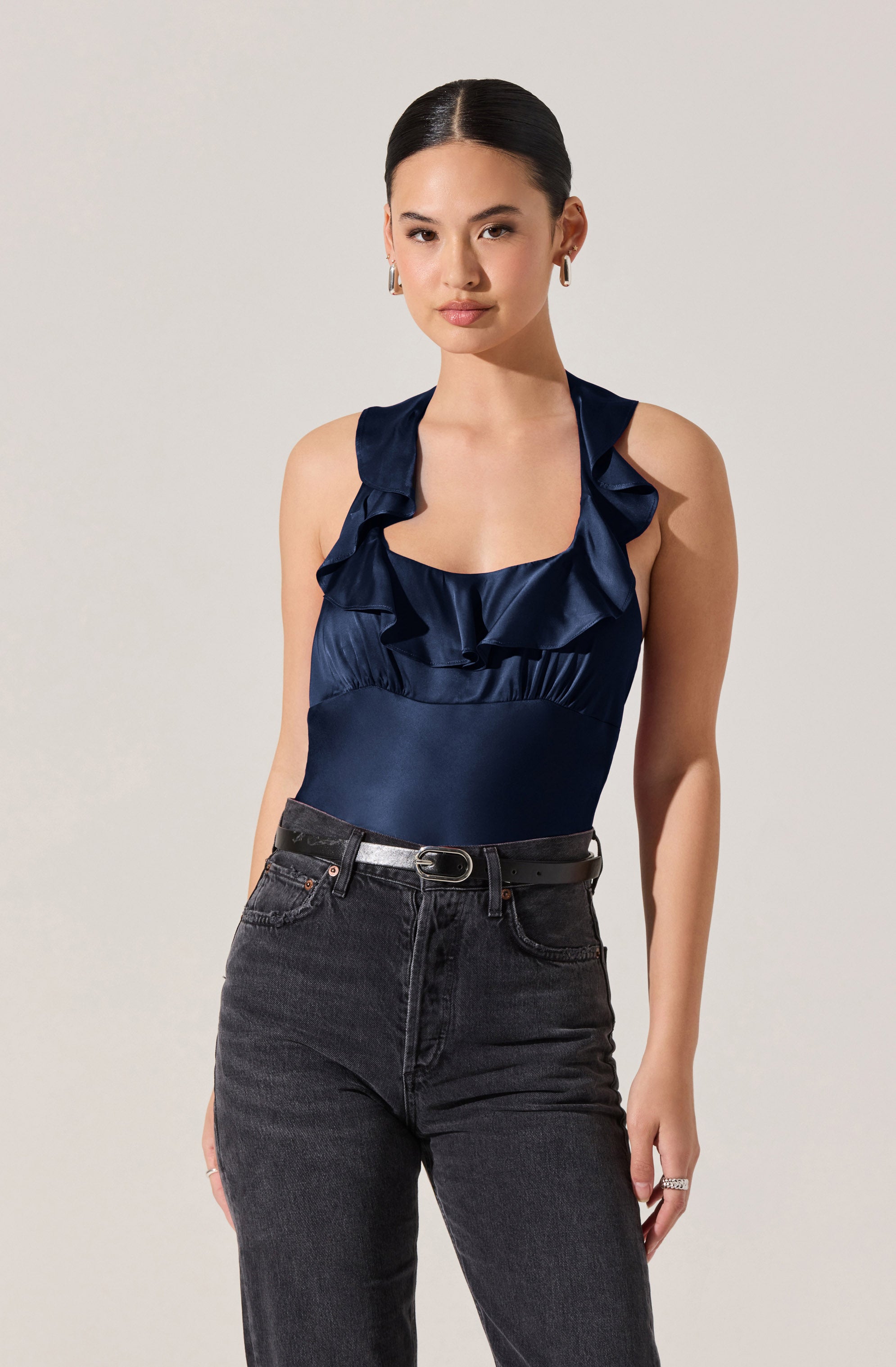 A woman is wearing a sleeveless navy blue satin top with ruffle details along the shoulders and neckline. She has paired it with high-waisted black jeans and a black belt with a silver buckle.