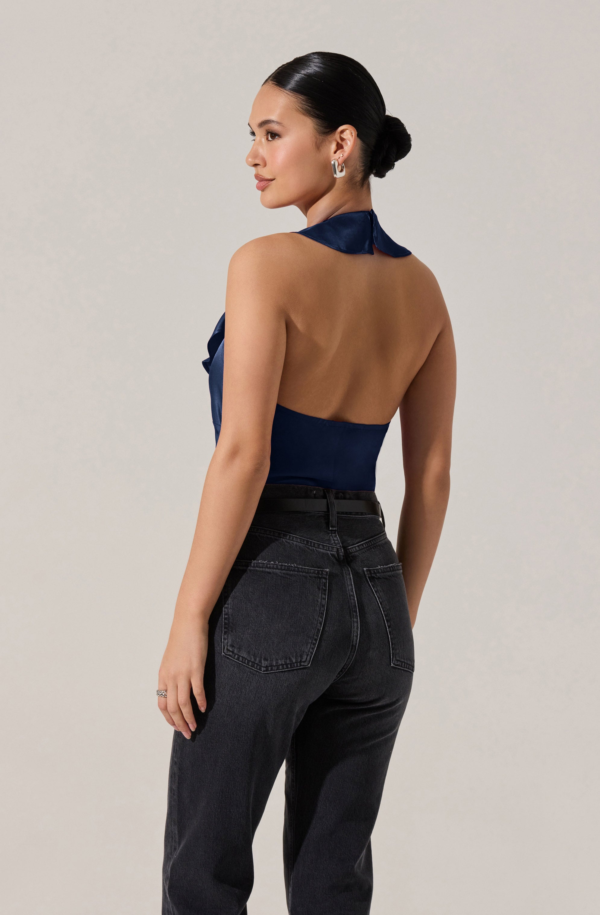 A woman is wearing a sleeveless, backless navy blue top with a ruffled halter neck, paired with high-waisted black jeans. The top has a shiny, satin-like fabric, and the jeans have visible stitching on the back pockets. The woman's hair is styled in a low bun, and she is accessorized with hoop earrings and a ring on her left hand.