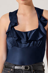A woman is wearing a sleeveless navy blue satin top with ruffled details around the neckline and shoulders. The top is fitted at the waist. She has paired it with black pants and a black belt with a silver buckle.
