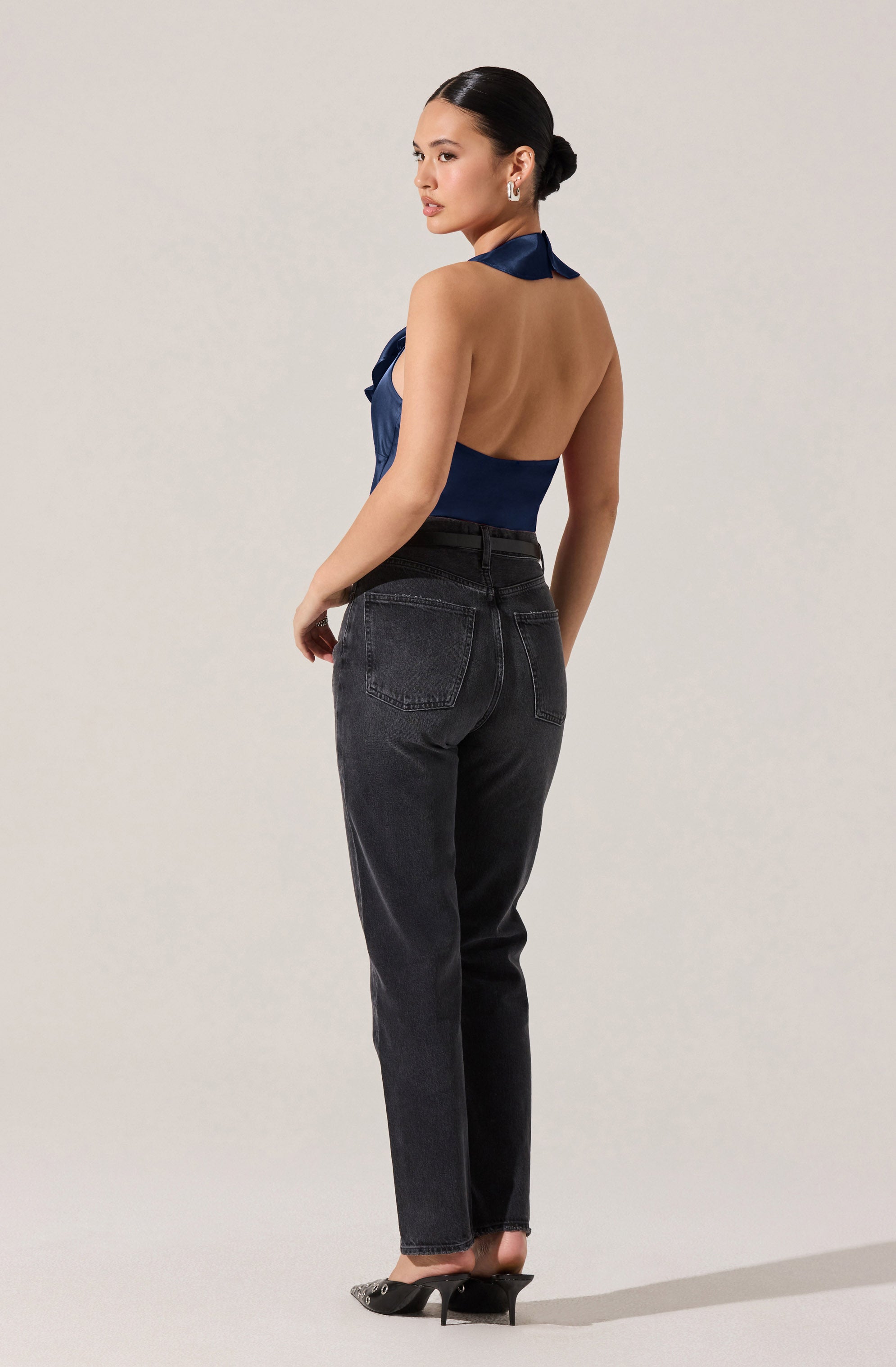 A woman is wearing a sleeveless, halter-style top in a shiny navy blue fabric. The top is backless and tied around her neck, exposing her back. She pairs it with high-waisted, dark-washed jeans and black high-heeled shoes.