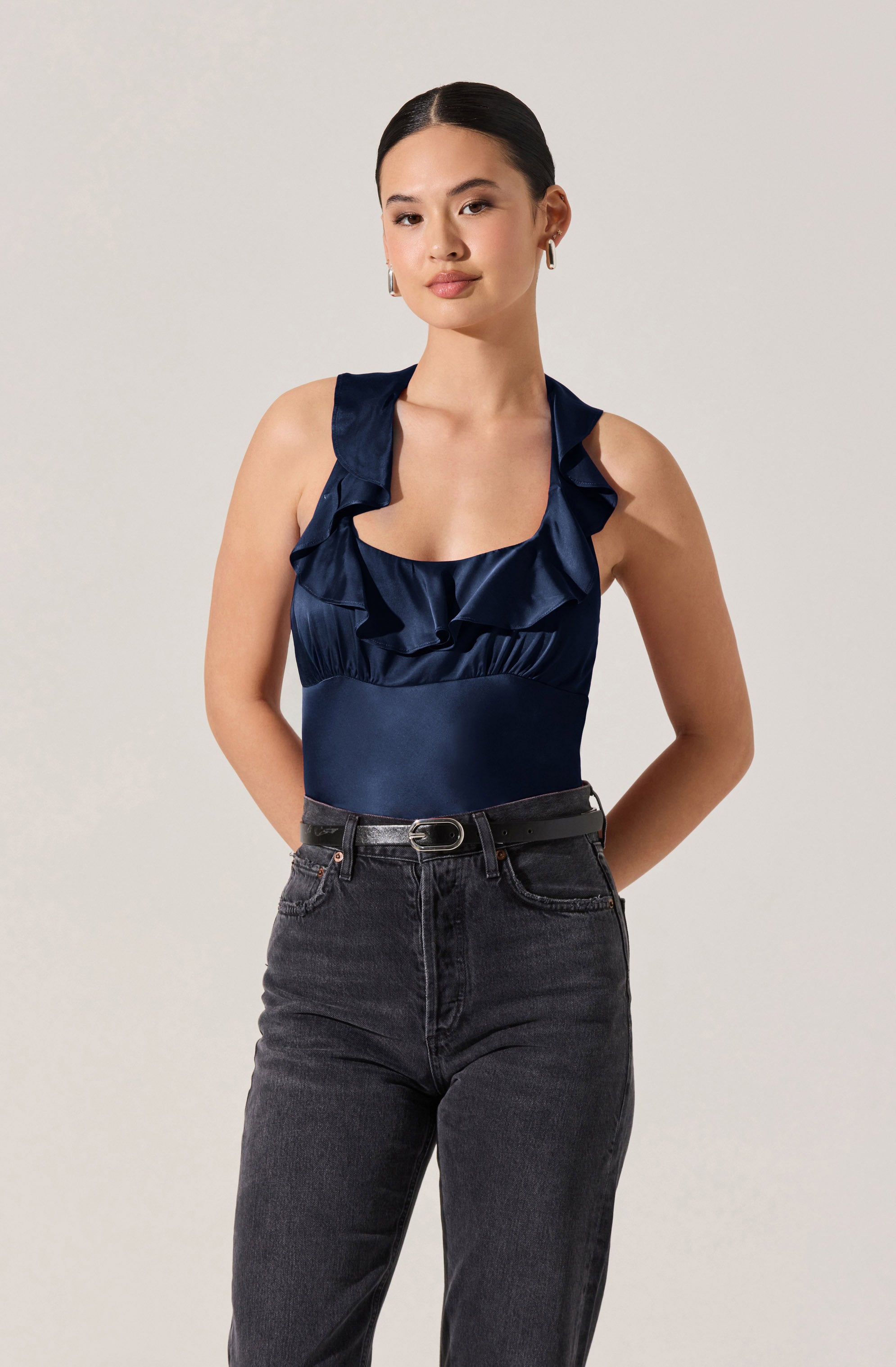 A woman is wearing a navy blue sleeveless top with ruffled shoulder straps and a ruffled neckline, made from a shiny satin-like fabric. She is also dressed in high-waisted, dark-wash jeans with a black belt.