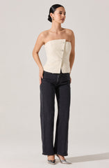 A woman is wearing a strapless, cream-colored, textured top with large buttons down the front. She pairs it with high-waisted, straight-leg black jeans and shiny silver pointed-toe heels. Her hair is styled in a sleek, low bun, and she has on gold earrings.