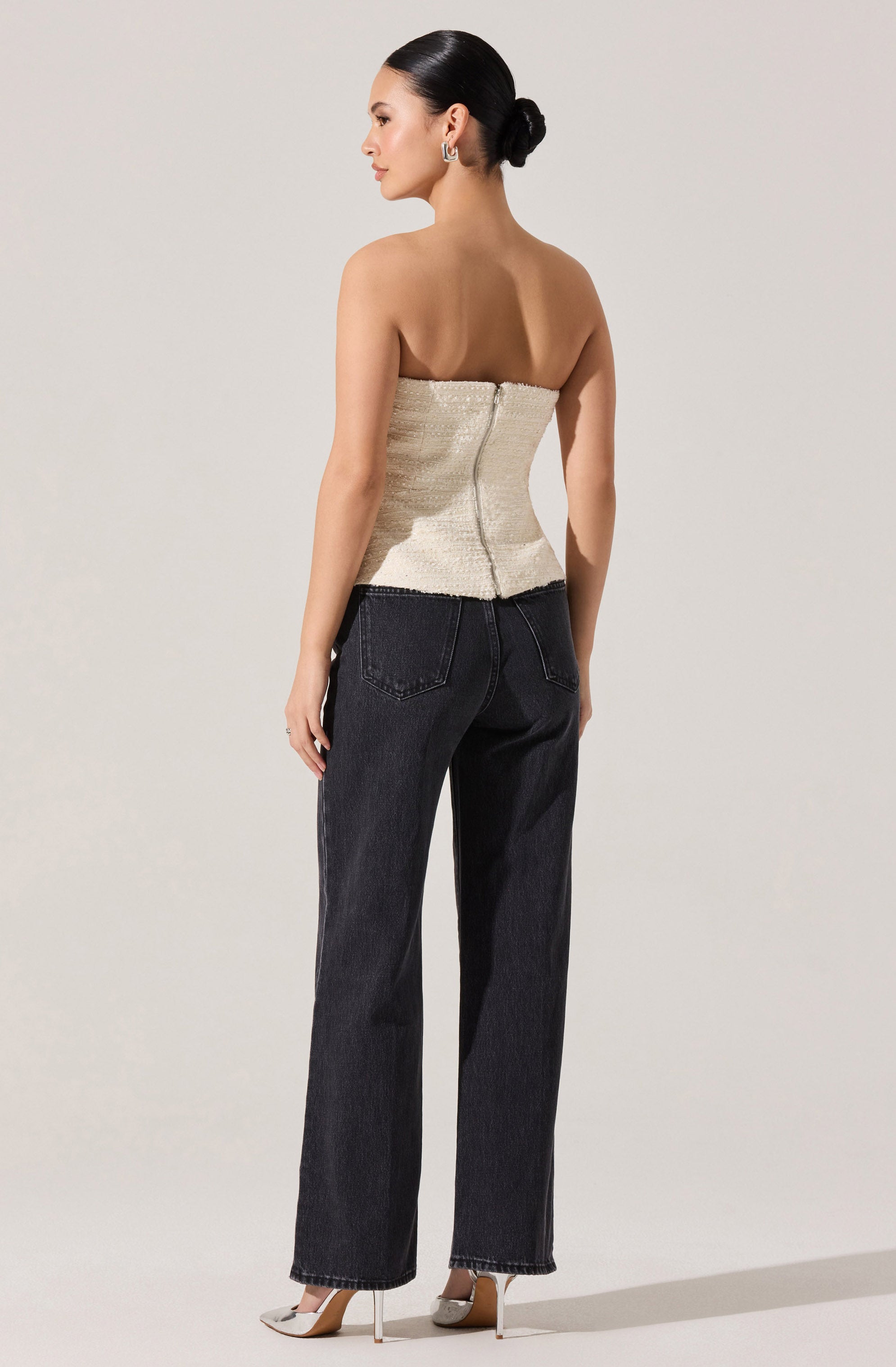 A woman is wearing a strapless, textured cream-colored top that is fitted and extends slightly below the waist, with a visible zipper at the back. She pairs it with high-waisted, straight-leg black jeans that have classic five-pocket styling. She completes her outfit with pointy-toed high heels that are a light neutral color.