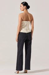 A woman is wearing a strapless, textured cream-colored top that is fitted and extends slightly below the waist, with a visible zipper at the back. She pairs it with high-waisted, straight-leg black jeans that have classic five-pocket styling. She completes her outfit with pointy-toed high heels that are a light neutral color.