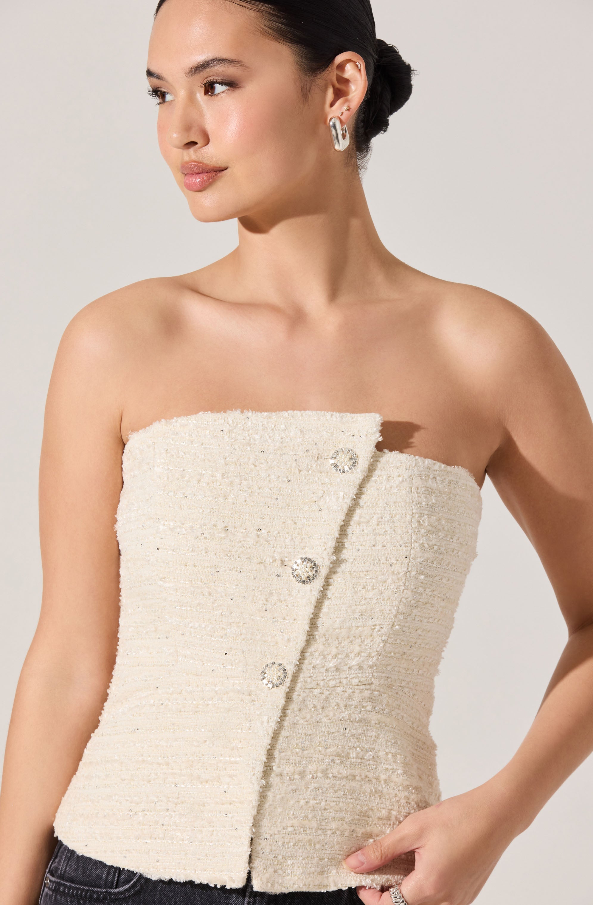 A woman is wearing a sleeveless, off-the-shoulder cream-colored top made of textured, woven fabric. The top features a diagonal row of large, round, decorative buttons with rhinestone embellishments. She has her dark hair styled in a bun and is accessorizing with silver hoop earrings.
