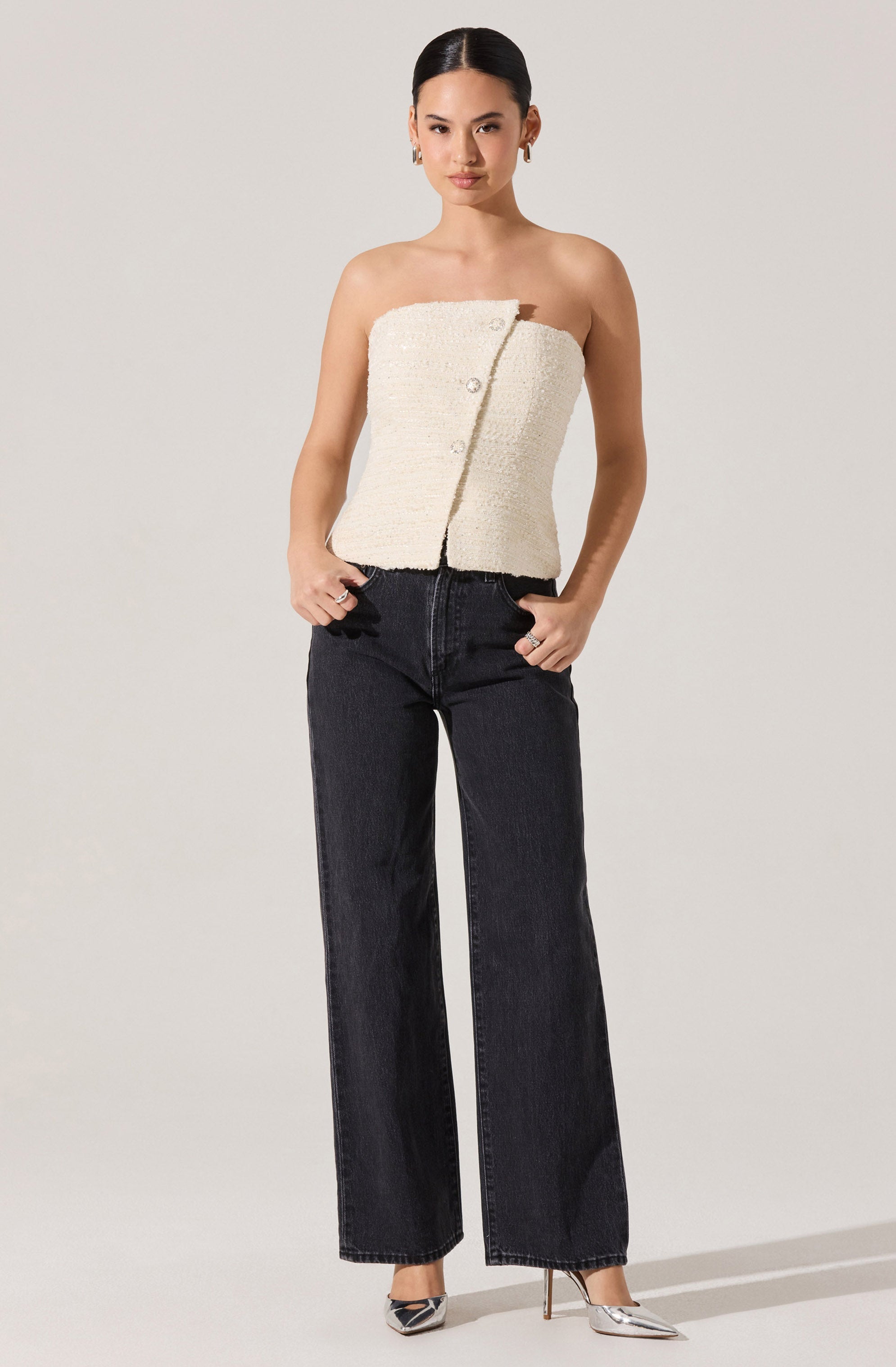 A woman is wearing a strapless, textured, off-white top with decorative buttons on the front. She pairs it with high-waisted, wide-leg black jeans and silver pointed-toe high heels.