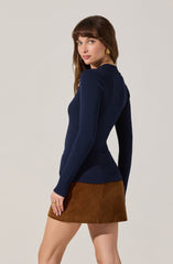 A woman is wearing a fitted, long-sleeved navy blue ribbed sweater with a high neckline and a brown corduroy mini skirt. She has gold hoop earrings and a gold ring on her right hand.