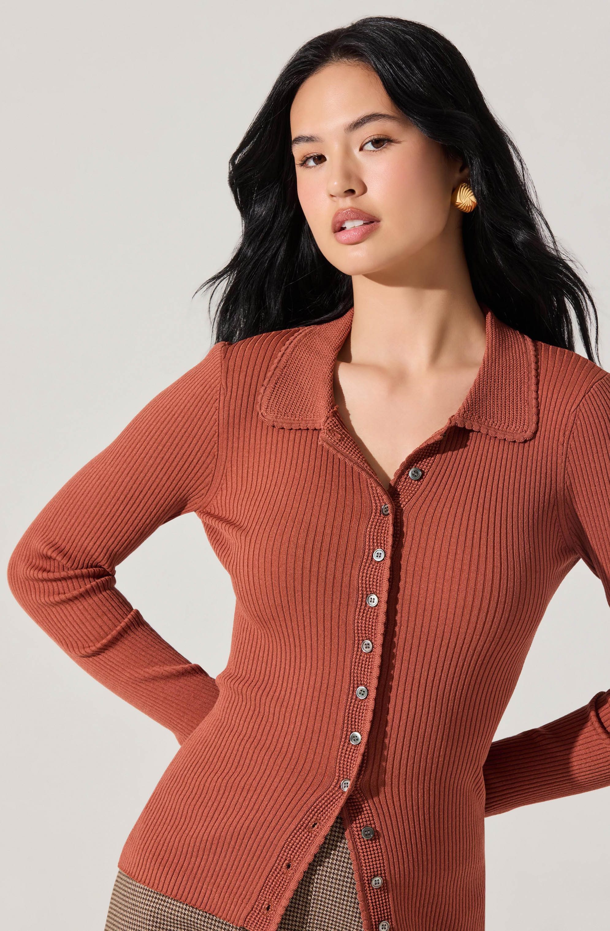 A woman is wearing a fitted, long-sleeve, ribbed cardigan in a warm rust color with a collar and buttons down the front. She has her hands on her hips and is accessorized with large, gold, shell-shaped earrings.