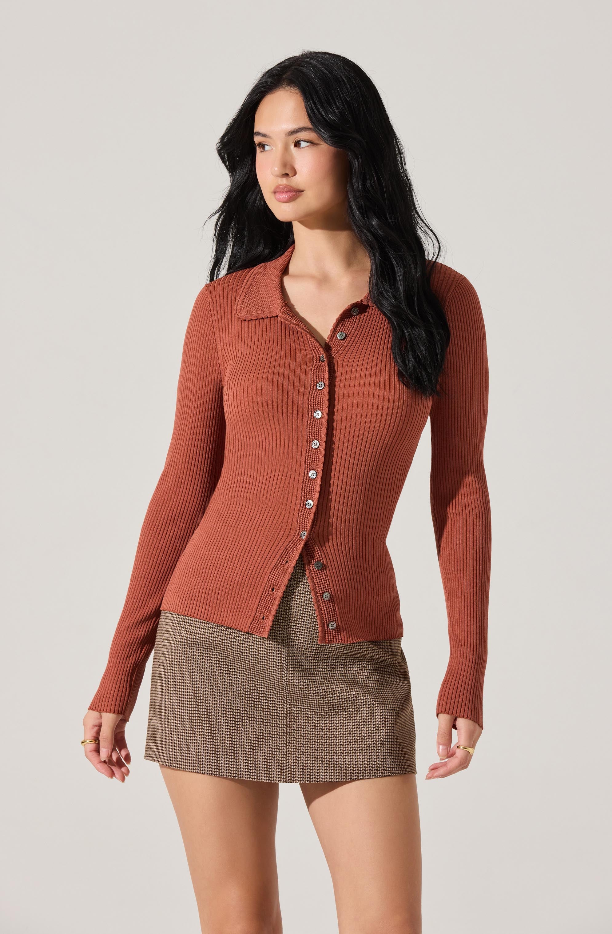 A woman is wearing a fitted, long-sleeve, ribbed, burnt orange cardigan with a collar and a row of buttons down the front. She is also dressed in a high-waisted, mini skirt with a small checkered pattern in neutral tones.